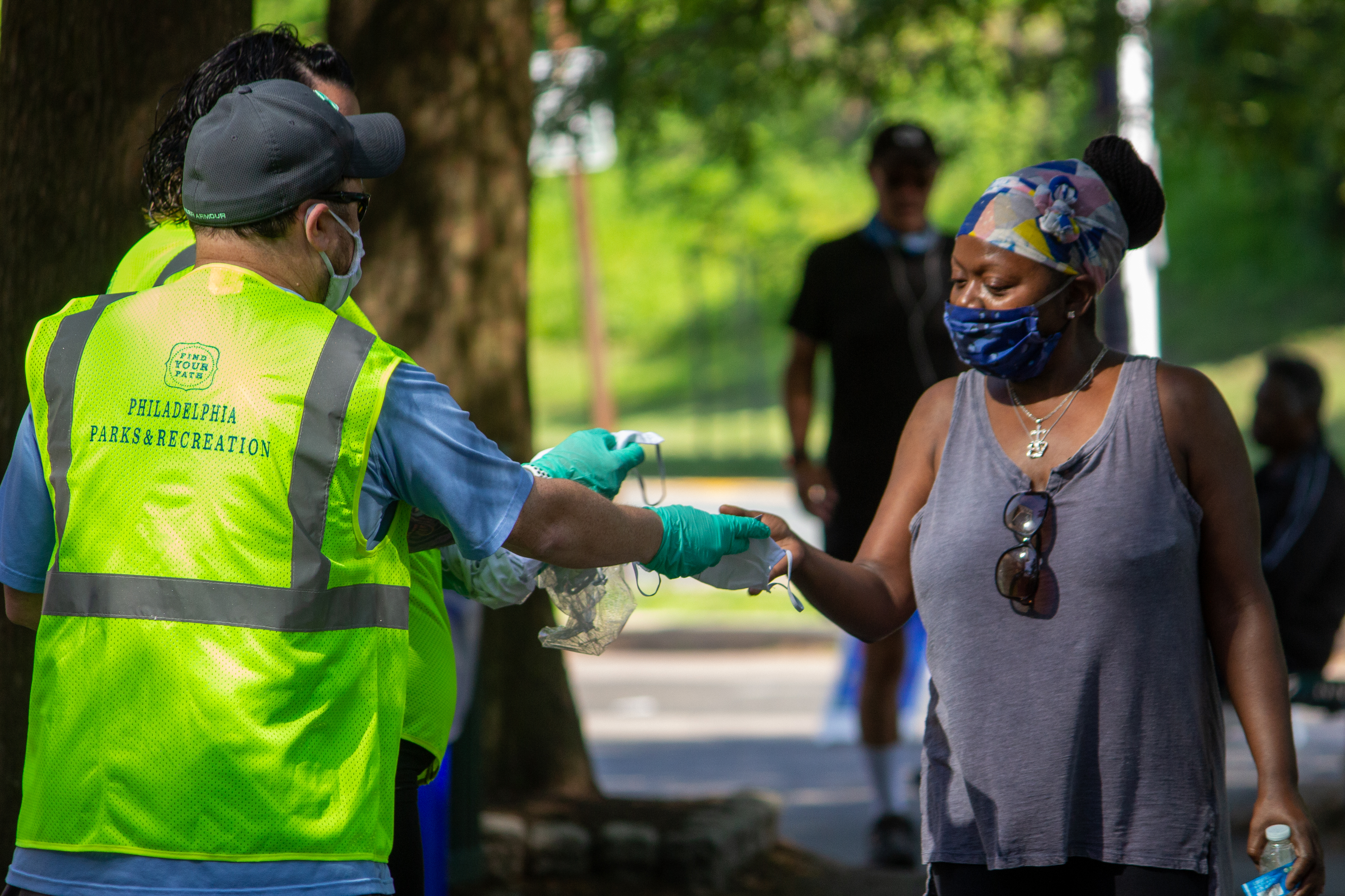 A man in a yellow Philadelphia Parks & Recreation vest handing a mask to a woman in a park. 