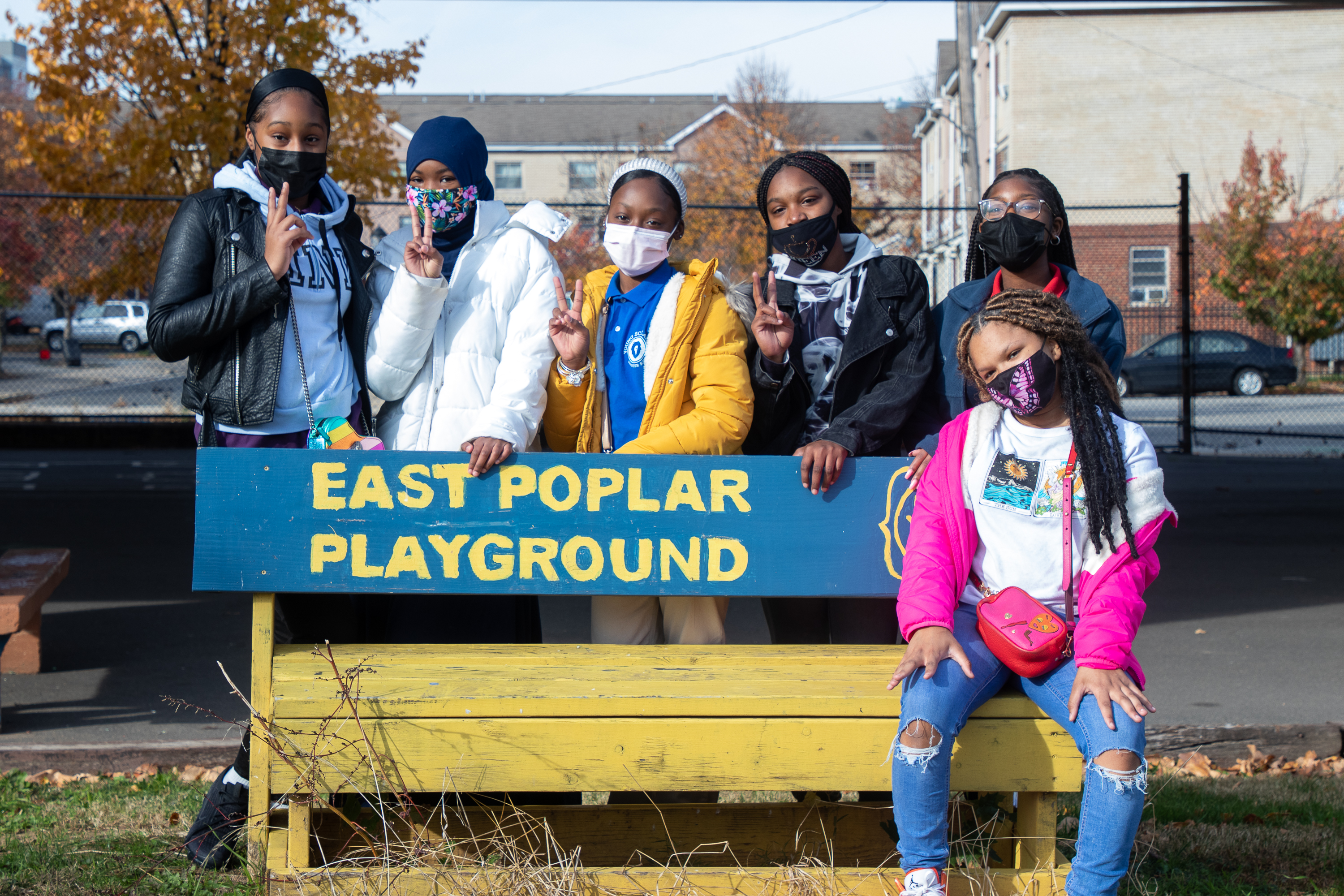 A girl sitting on a blue and yellow bench that says East Poplar Playground and five girls standing behind the bench.