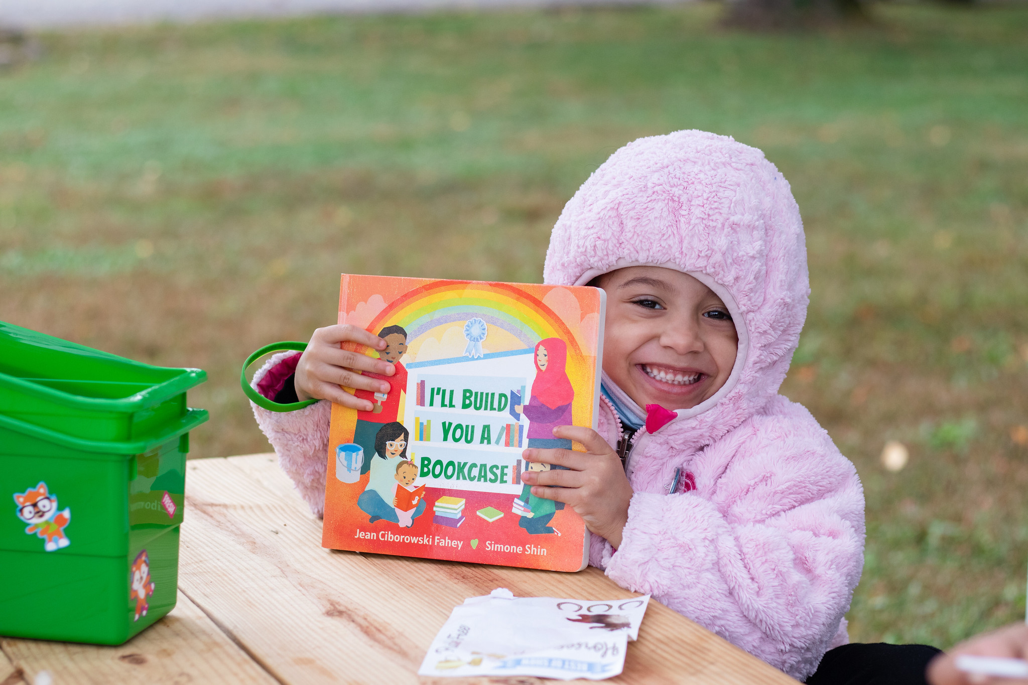 A young girl in a fuzzy pink jacket with the hood up, sitting at a picnic table, holding a book.