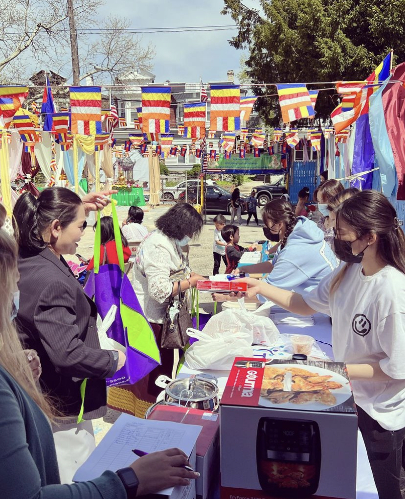 A group of people outside, on opposite sides of a table with colorful flag garlands overhead.