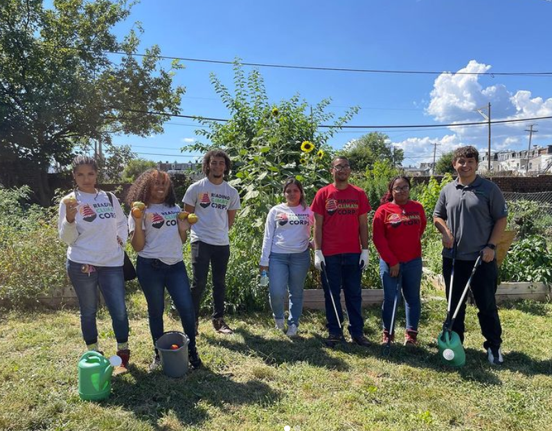 A group of 7 people standing in a community garden holding squash and garden tools