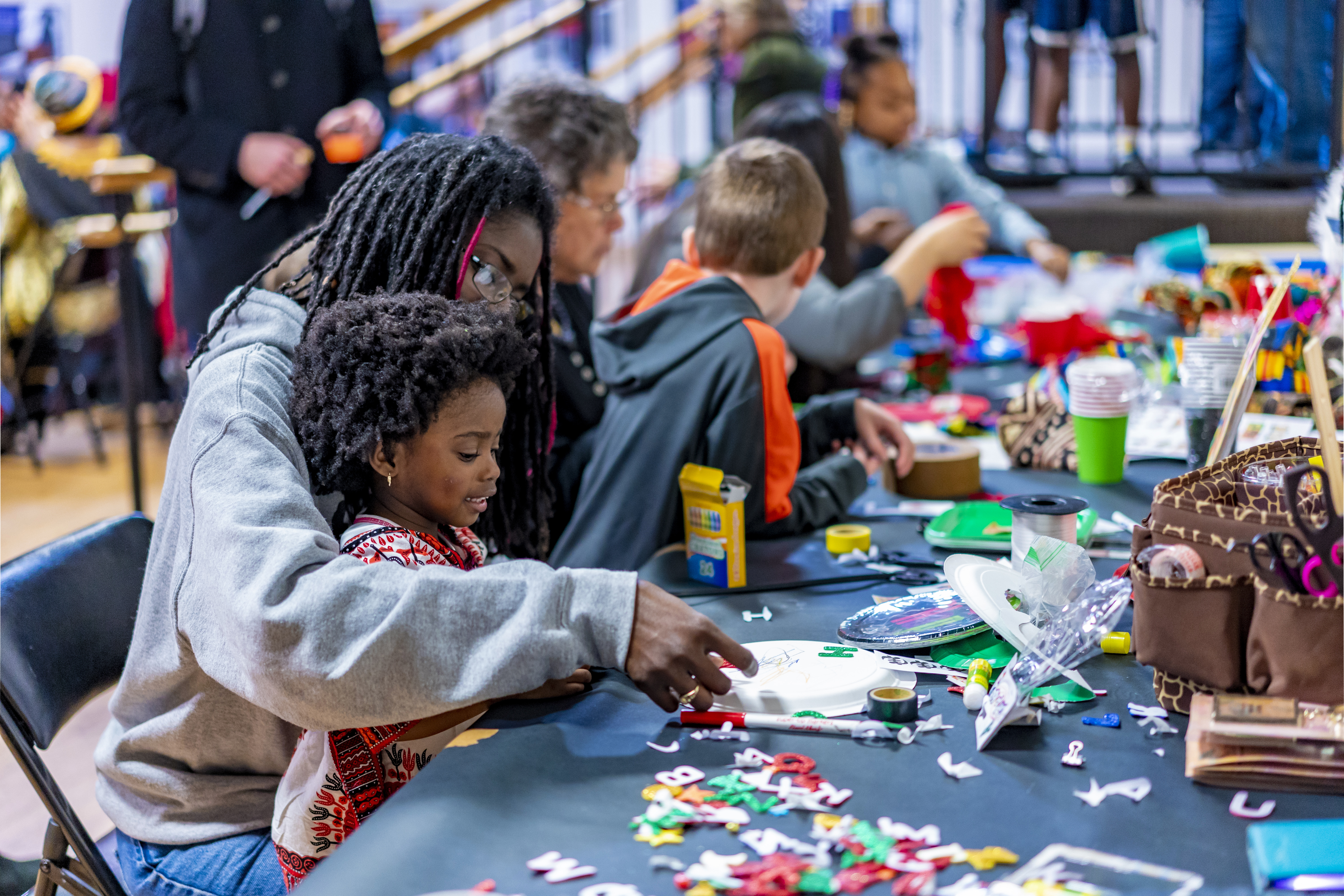 A woman and child seated at a long table covered with craft supplies, working on a project. 