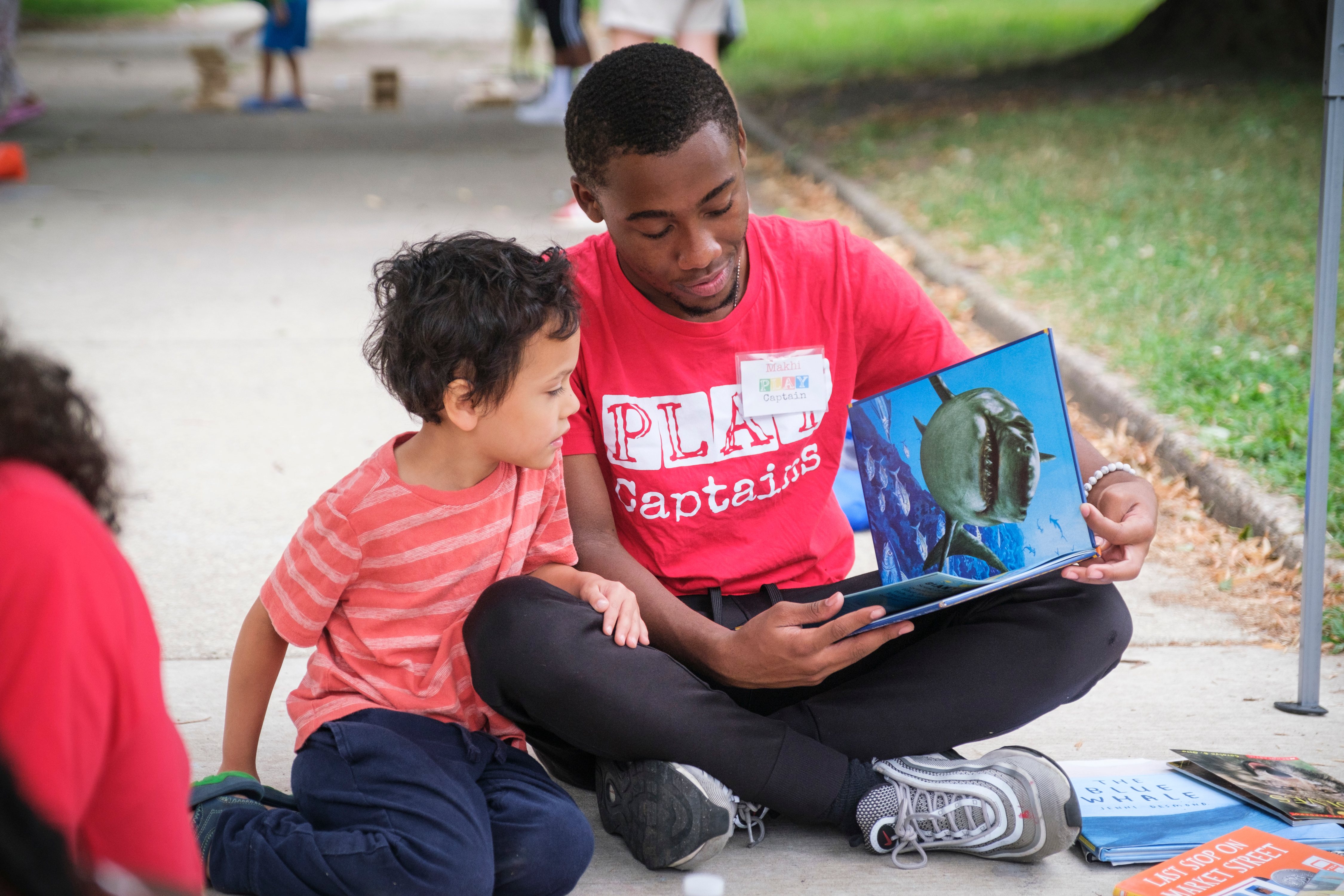 A young man sitting on the ground reading a book to a child.