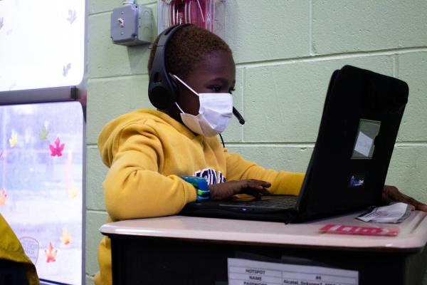 A child wearing a mask, sitting at a desk working on a laptop